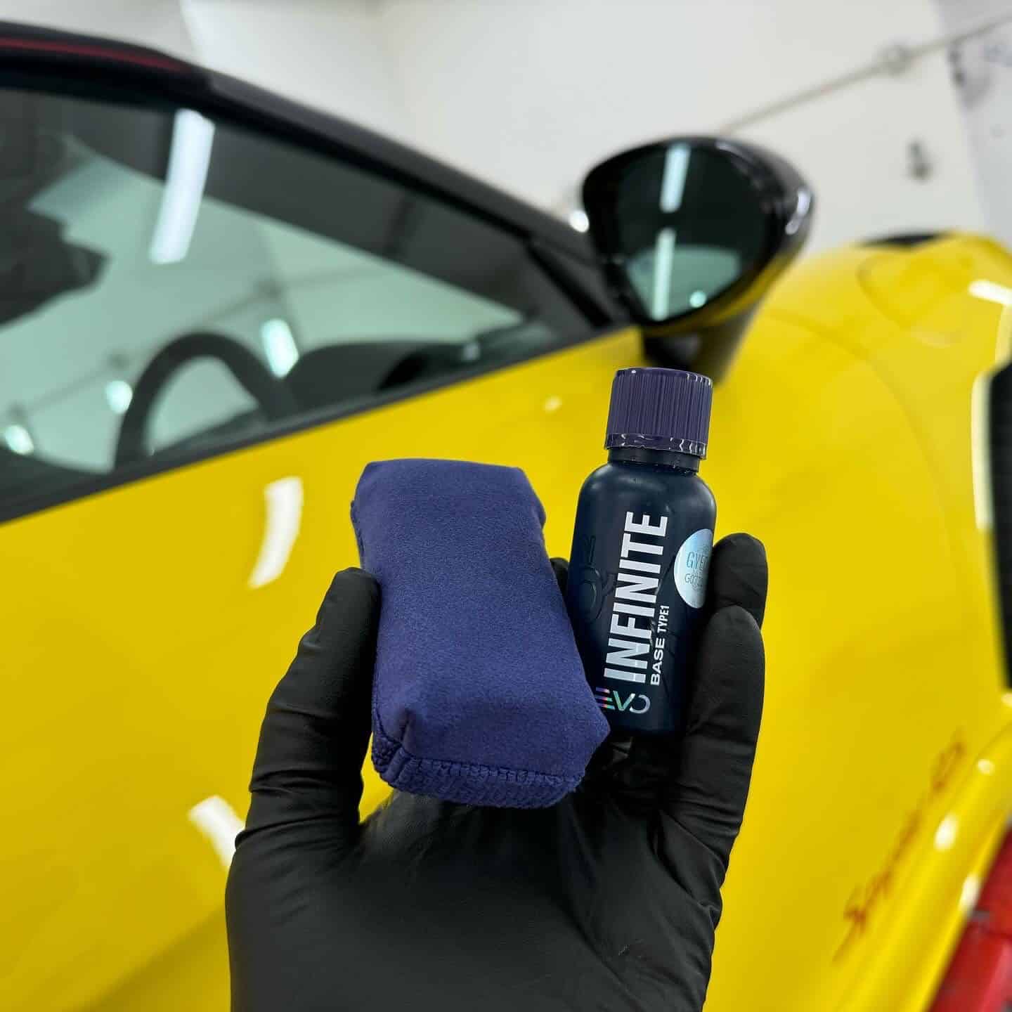 Holding a Gyeon ceramic coating bottle and applicator, with a Porsche 718 Spyder RS in the background
